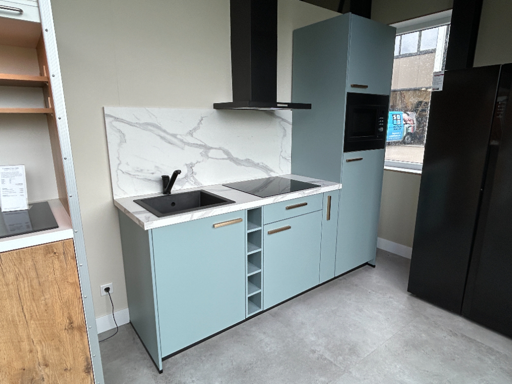kitchenatwork Een moderne keuken met lichtblauwe kasten, wit aanrecht, zwarte spoelbak, kookplaat, oven en een achterwand met marmerpatroon. Boven de kookplaat bevindt zich een ventilatiekap. Op de achtergrond is een venster zichtbaar.